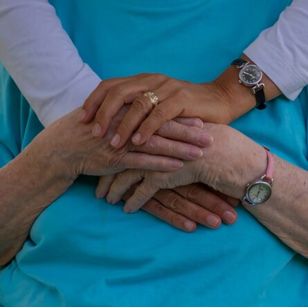 A close-up of two pairs of hands, one older and one younger, gently resting on top of each other, symbolizing compassion