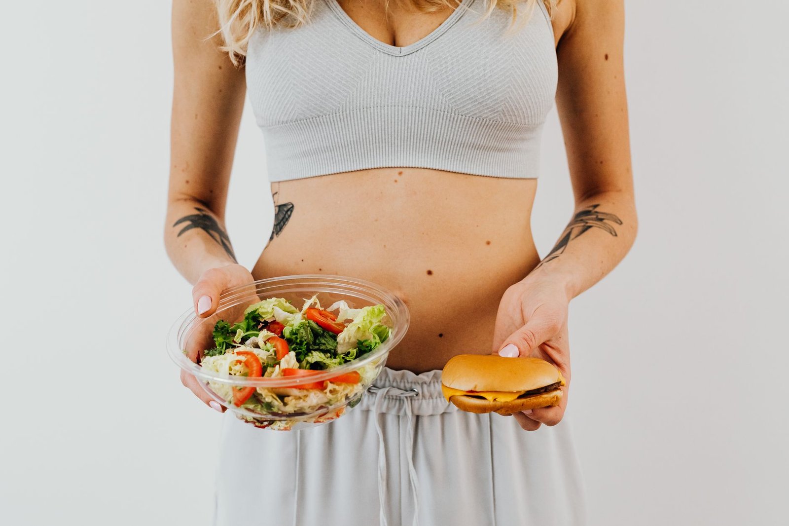 A woman in a sports bra holding a salad and cheeseburger, representing healthy vs junk food choices. — 3 of the Most Effectiv