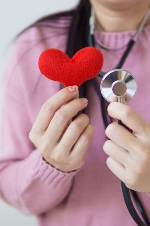 Unrecognizable female in pink sweater with stethoscope on neck standing on white background with red heart in hand in dayligh