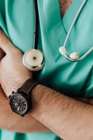 Unrecognizable man wearing medical uniform with stethoscope and pens standing in hospital with crossed arms during work in da
