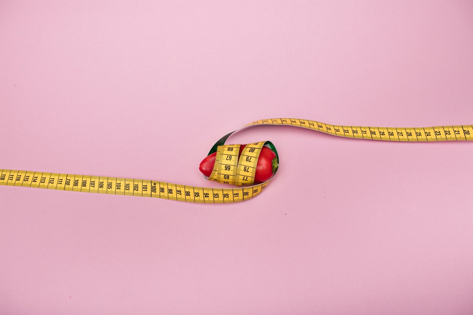 A red bell pepper wrapped with a yellow measuring tape on a pink background symbolizing diet and health. — Health Care Reform