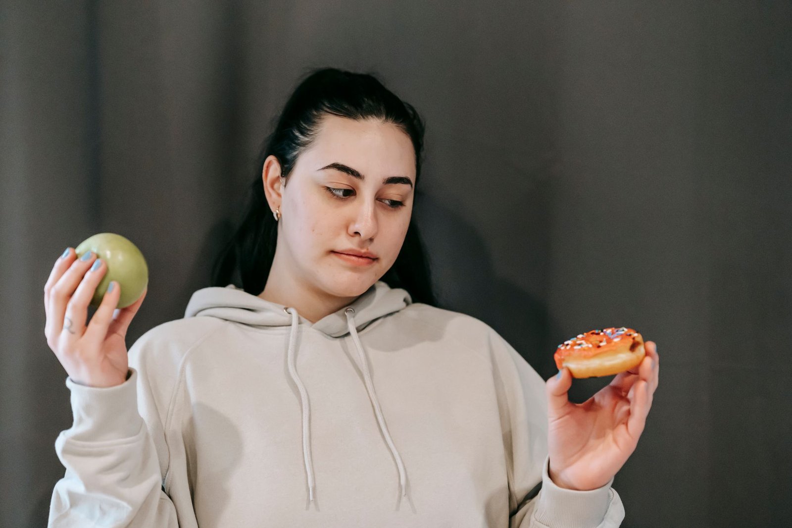 Contemplative plump female in hoodie choosing between ripe green apple and delicious donut in studio — Live Longer - 7 Truths