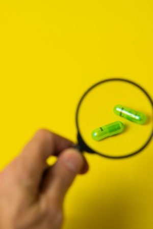From above of crop unrecognizable male holding magnifying glass over green pills on yellow background in studio — Smart Choic