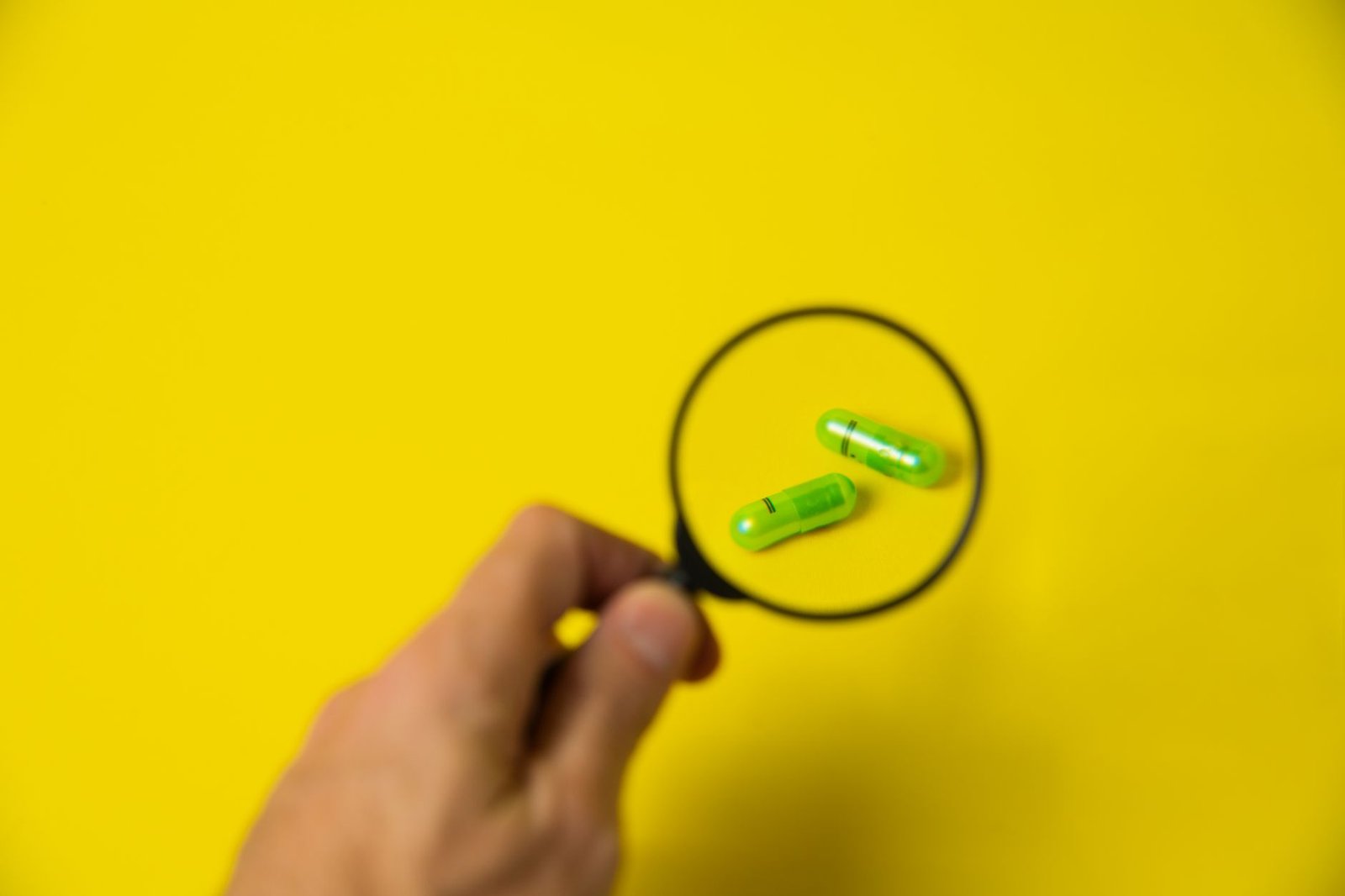 From above of crop unrecognizable male holding magnifying glass over green pills on yellow background in studio — Smart Choic