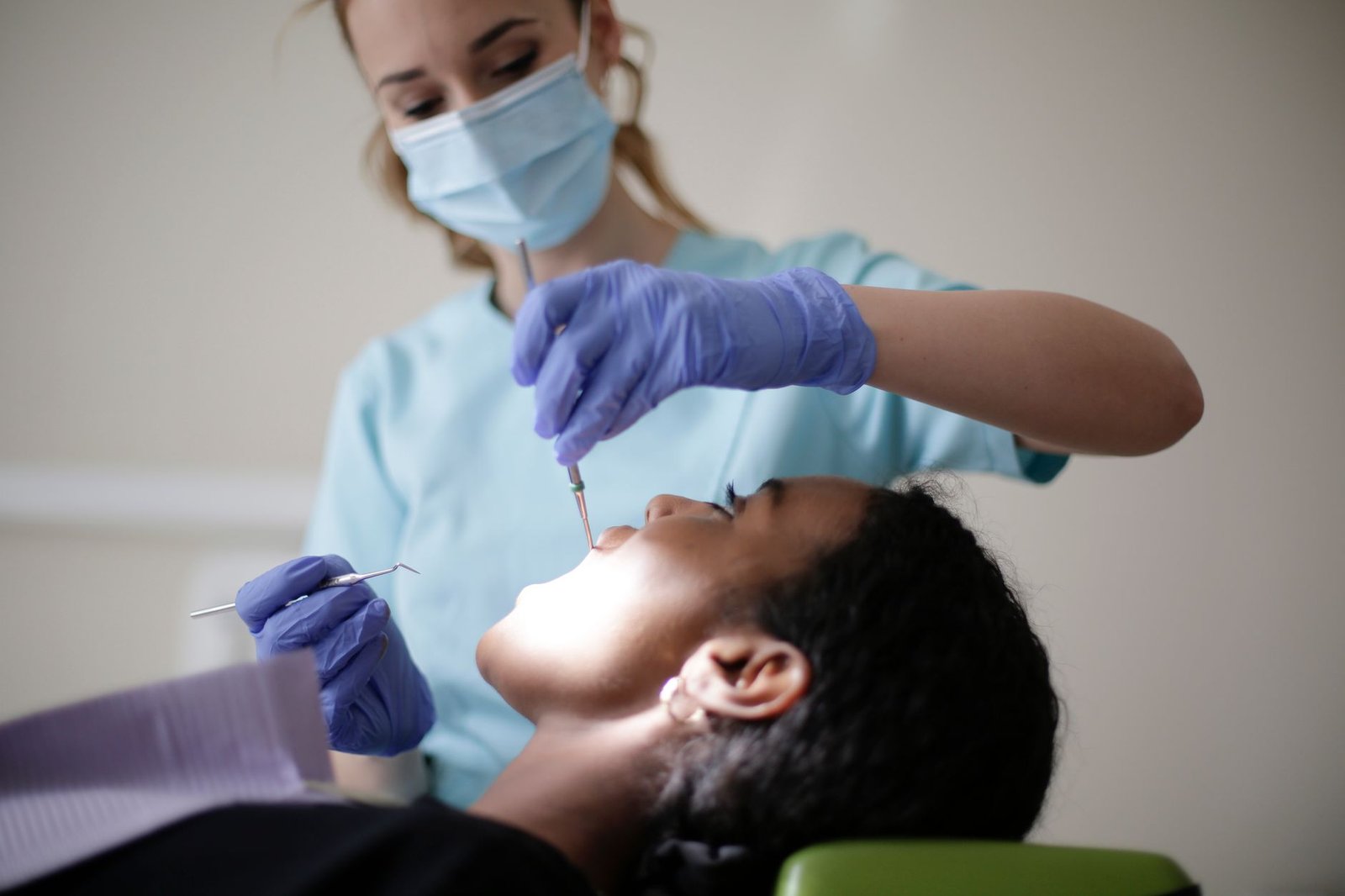 Serious dentist in latex gloves mask and uniform using stomatological instruments for examining teeth of African American pat